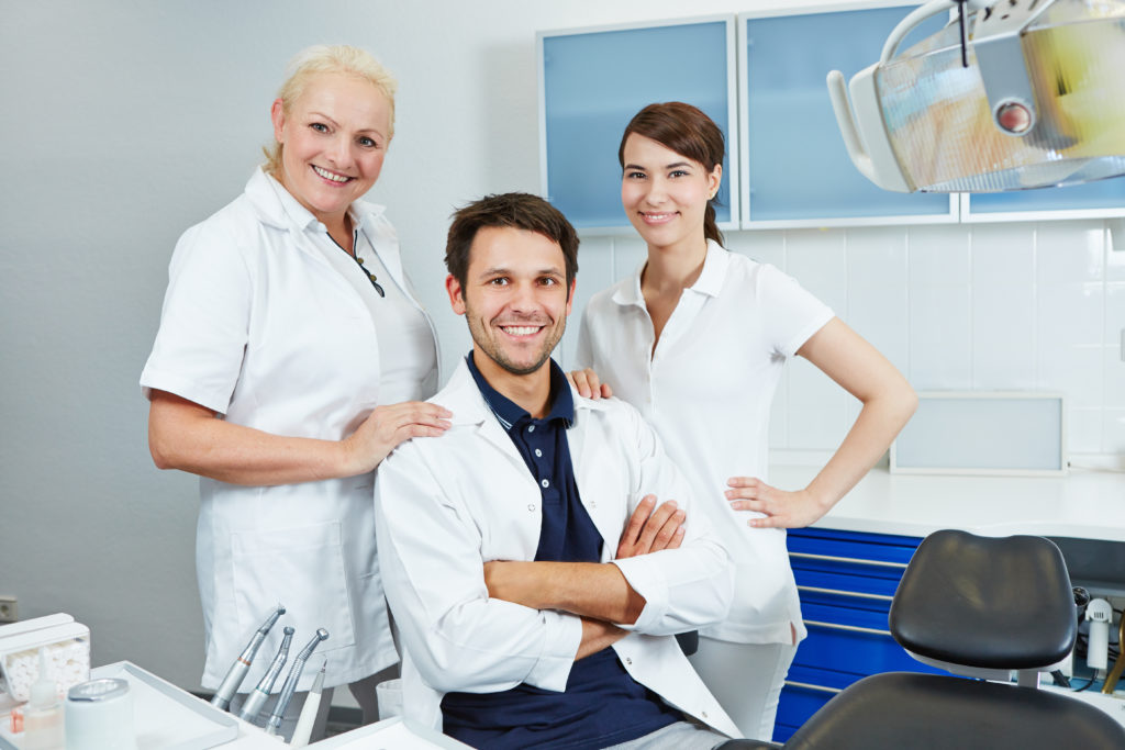 A dentist and two female dental assistants, all in white, smile in the patient area